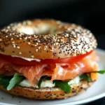 An everything bagel sandwich filled with cream cheese, smoked salmon, fresh tomato slices, and arugula on a white plate.