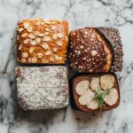 Four beautifully baked loaves of bread, each with unique textures and toppings, displayed on a marble background