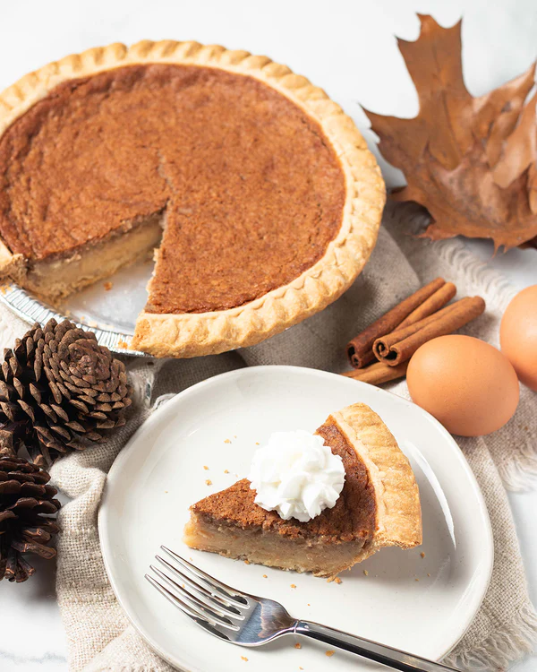 A freshly baked bean pie with a golden crust, served with a slice on a white plate, topped with whipped cream. Cinnamon sticks, eggs, pinecones, and autumn leaves are arranged around the pie, creating a cozy fall-themed setting.