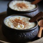 Two bowls of creamy rice pudding (Arroz con Leche) topped with cinnamon powder and garnished with cinnamon sticks, served in rustic clay bowls.